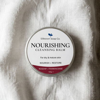 A round tin of Silktown Soap Co. Nourishing Cleansing Balm, featuring a white label with black text and a dark red bottom banner, set against a textured white linen background.