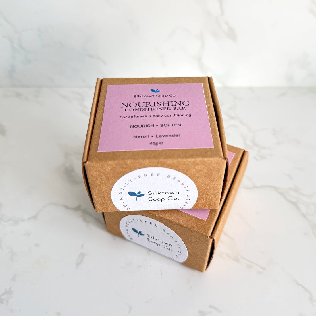 Two small kraft paper boxes of Silktown Soap Co. Nourishing Conditioner Bar stacked on a white marble surface against a white marble background.