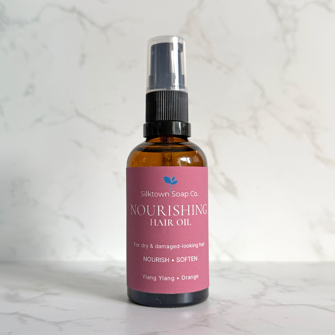A bottle of Silktown Soap Co. Nourishing Hair Oil in the Ylang + Orange scent, featuring an amber glass bottle with a pink label and a black spray nozzle, set against a white marble background.