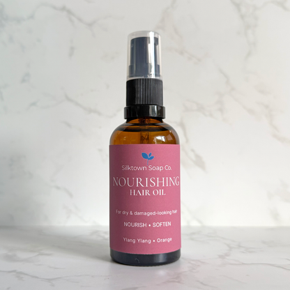 A bottle of Silktown Soap Co. Nourishing Hair Oil in the Ylang + Orange scent, featuring an amber glass bottle with a pink label and a black spray nozzle, set against a white marble background.