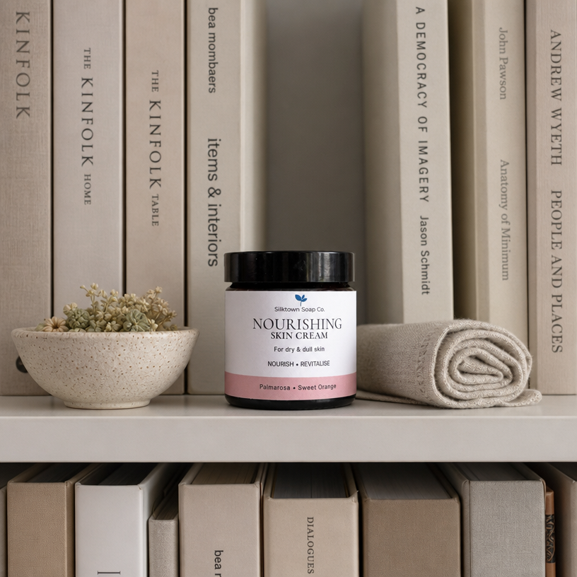 A jar of Silktown Soap Co. Skincare Nourishing Skin Cream sits on a white shelf styled with books, a small bowl of dried flowers, and a rolled beige towel.