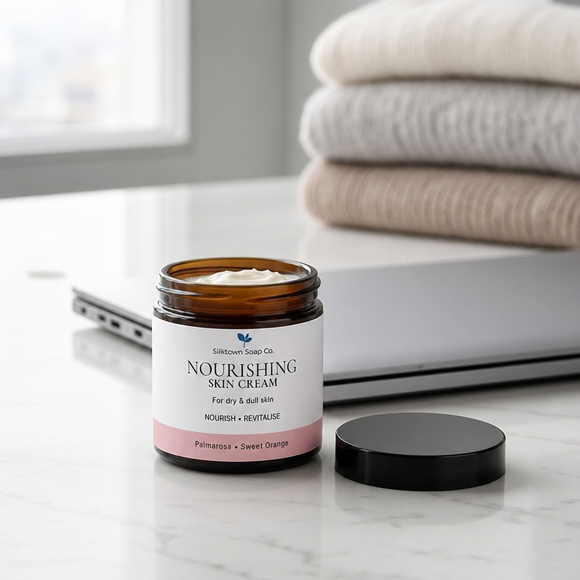 Silktown Soap Co. Nourishing Skin Cream in an amber glass jar with a white and pink label, shown with its black lid on a marble surface next to a laptop and folded towels.