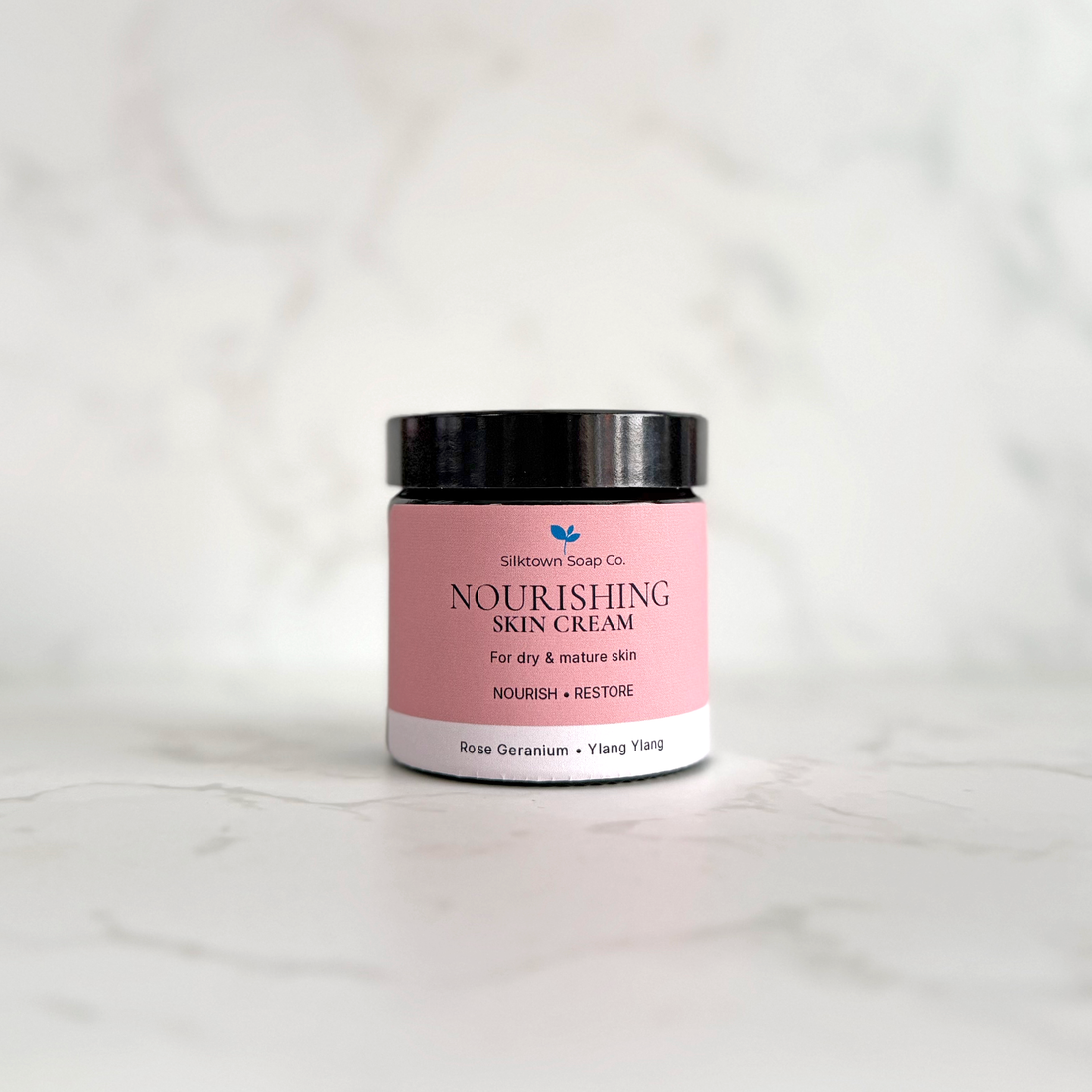A small cosmetic jar of Silktown Soap Co. Nourishing Skin Cream, featuring a pink label and black lid, set on a light gray marble surface against a matching marble background.
