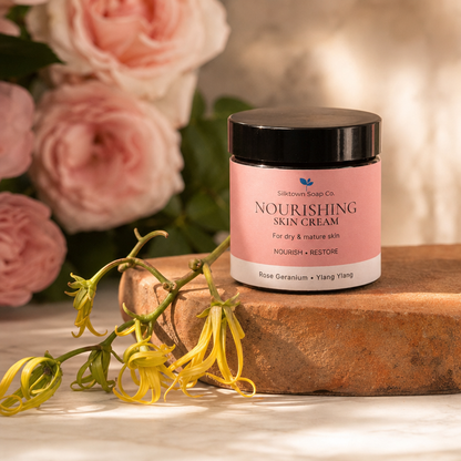 A jar of Silktown Soap Co. Nourishing Skin Cream sits on a rustic terracotta block, styled with fresh rose geranium and yellow flowers against a soft-focus background of pink roses.