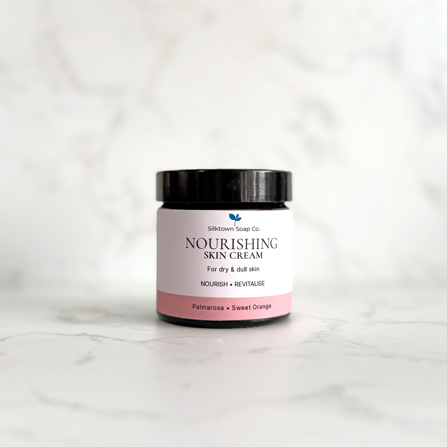 A small black jar of Siltown Soap Co. Nourishing Skin Cream with a white and pink label, set on a light gray marble surface against a matching marble background.