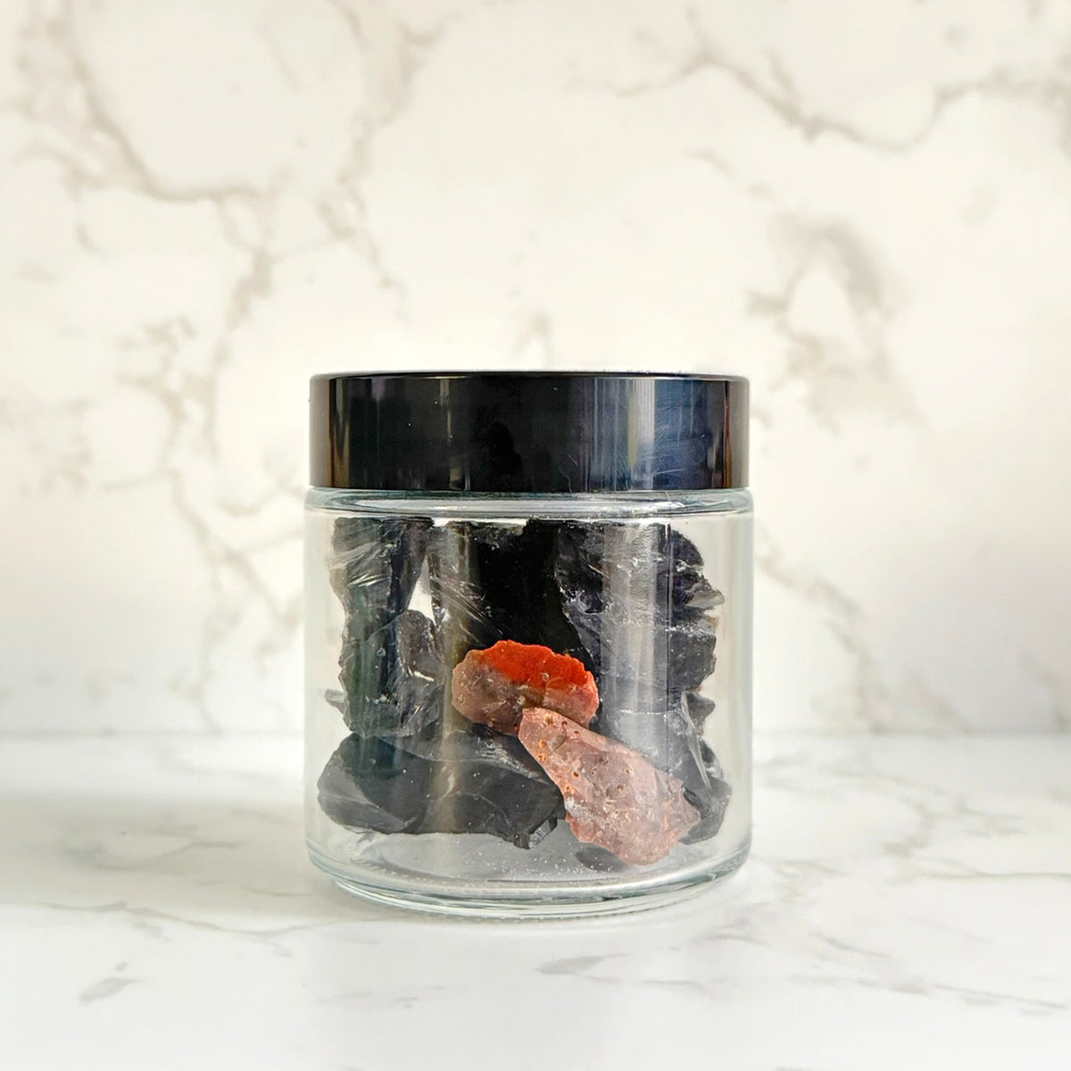 A clear glass jar with a black lid containing dark polished stones and two pinkish-orange raw crystal pieces, set on a white marble surface against a white marble background.