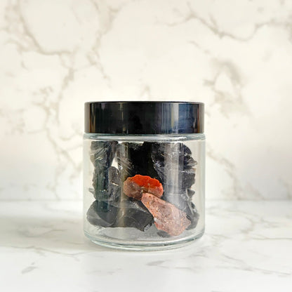 A clear glass jar with a black lid containing dark polished stones and two pinkish-orange raw crystal pieces, set on a white marble surface against a white marble background.