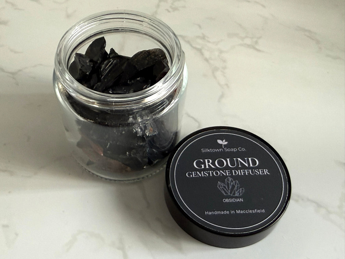 A clear glass jar filled with dark crushed gemstone pieces sits on a white marble surface next to a round black lid labeled &