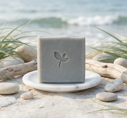 ocean breeze soap bar on white dish on sand with driftwood and pebbles with waves in the background
