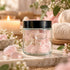 A clear glass jar with a black lid filled with polished pink rose quartz crystals, styled on a marble surface with a lit candle, rolled towels, and small flowers in the background.