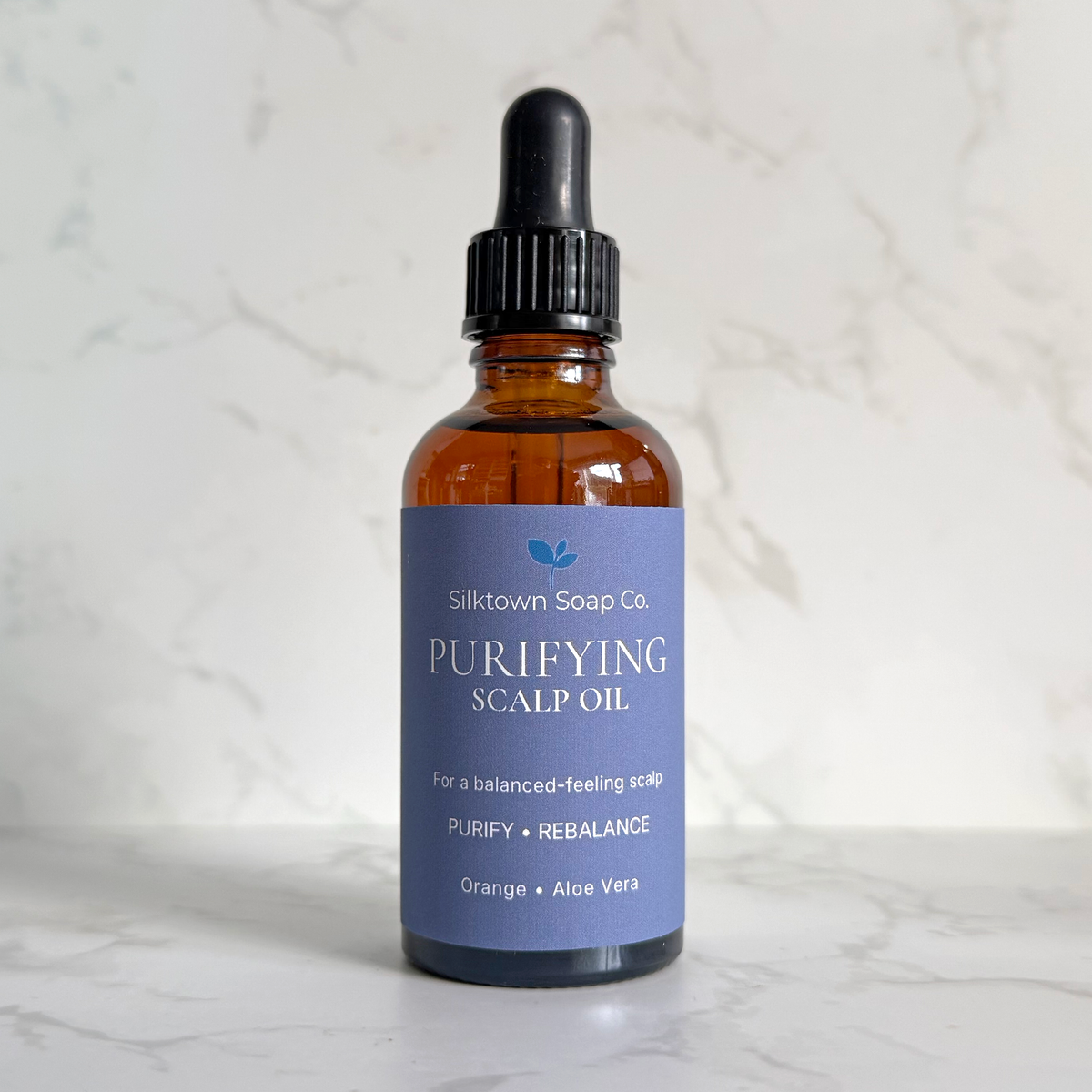 A bottle of Silktown Soap Co. Purifying Scalp Oil with an amber glass bottle, black dropper cap, and a lavender-blue label, set on a white marble surface against a white marble background.
