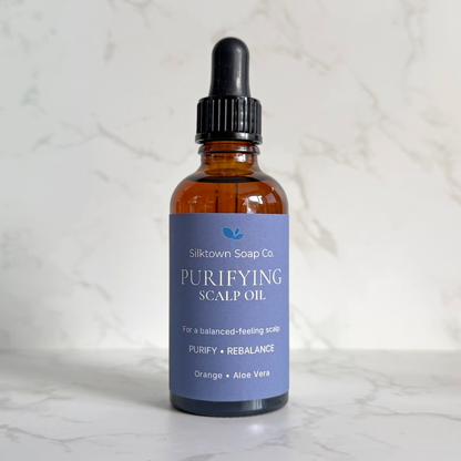 A bottle of Silktown Soap Co. Purifying Scalp Oil with an amber glass bottle, black dropper cap, and a lavender-blue label, set on a white marble surface against a white marble background.