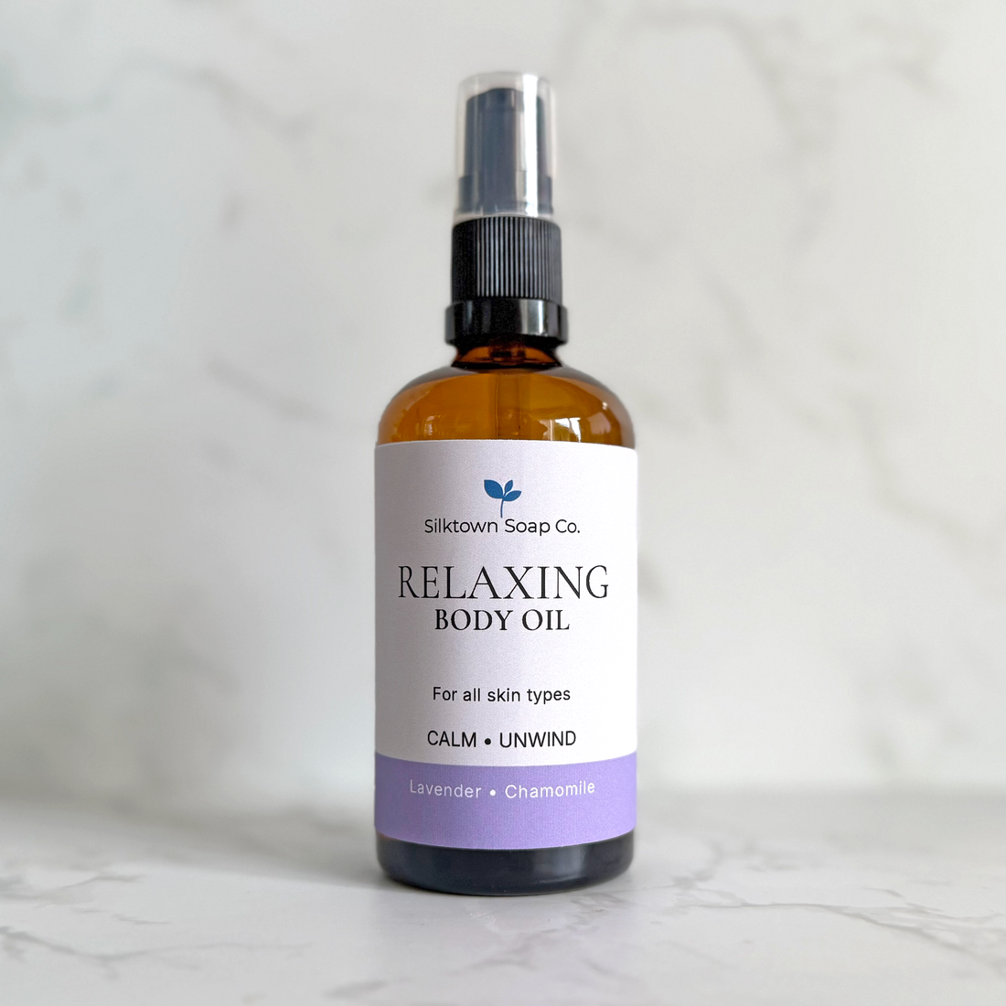 An amber glass spray bottle of Silktown Soap Co. Relaxing Body Oil with a white label and lavender accent, set against a light marble background.