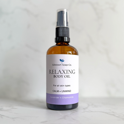 An amber glass spray bottle of Silktown Soap Co. Relaxing Body Oil with a white label and lavender accent, set against a light marble background.