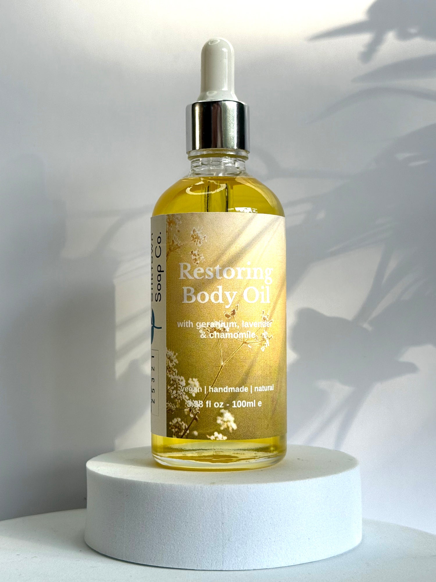 Bottle of Restoring Body Oil on a white pedestal with a white background