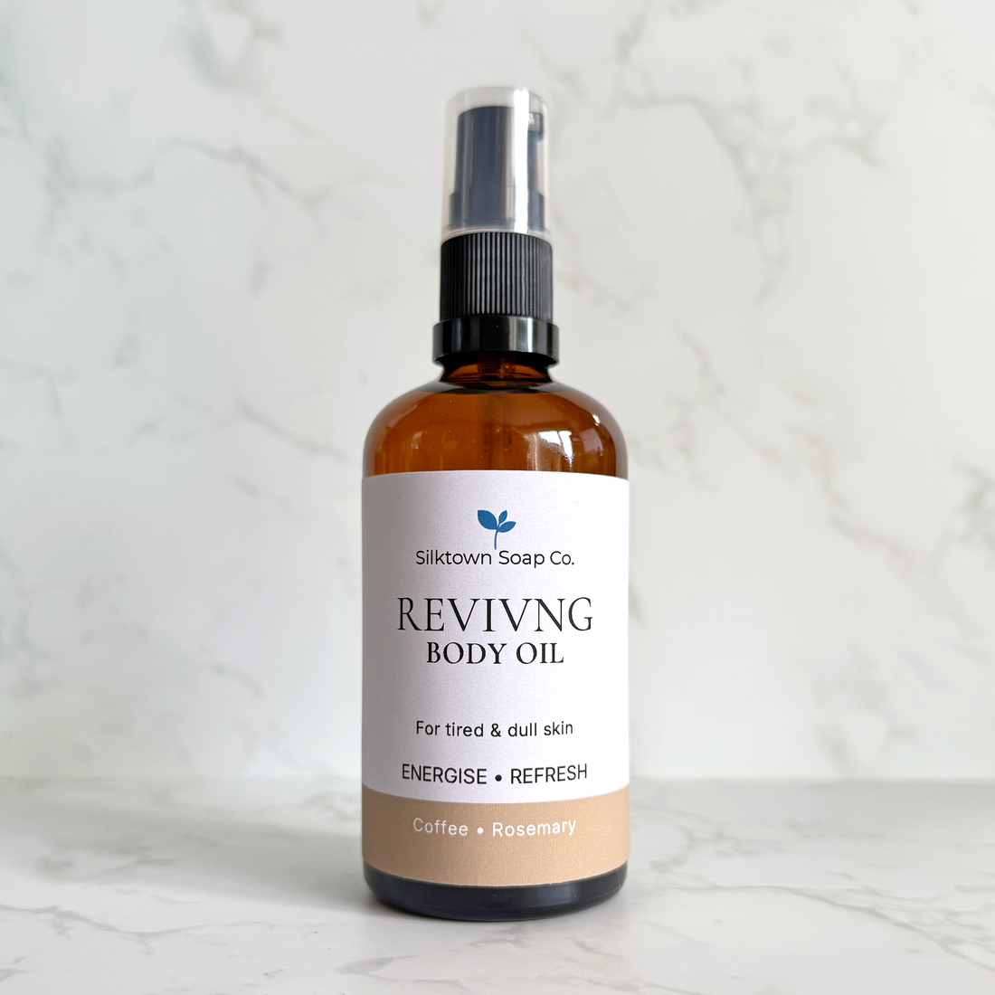 A bottle of Silktown Soap Co. Reviving Body Oil in a coffee and rosemary scent, featuring an amber glass bottle, black spray pump, and a white label with a beige bottom section, set against a light marble background.