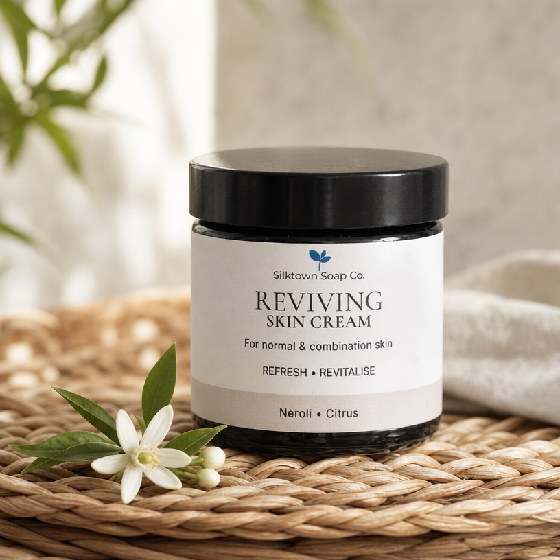 Silktown Soap Co. Reviving Skin Cream in a black jar with a white label, styled on a woven straw mat with a neroli flower.
