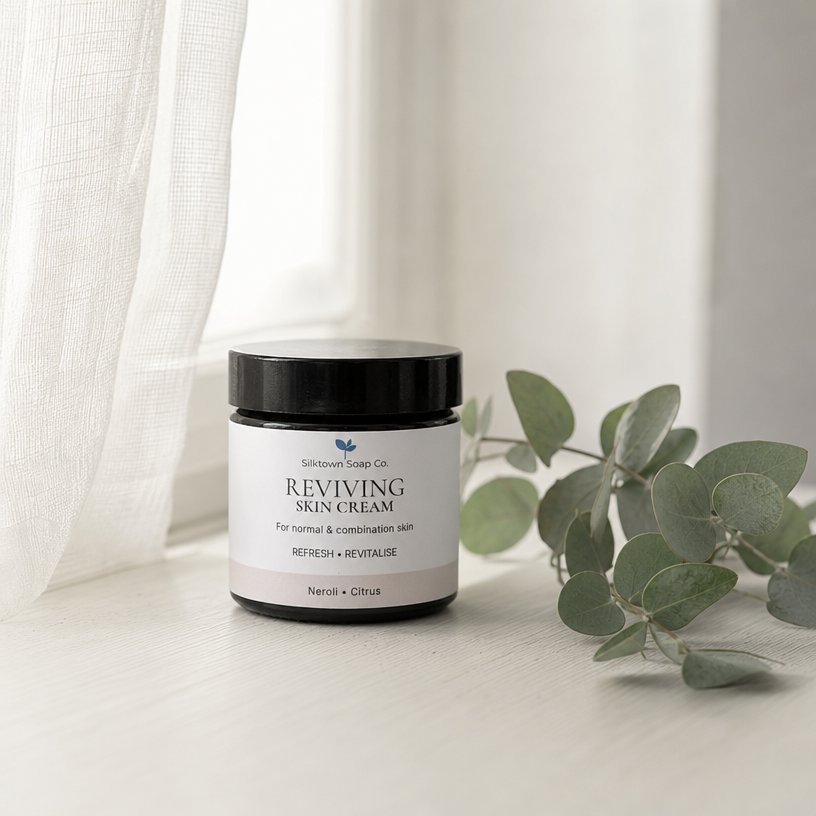 A jar of Reviving Skin Cream by Silktown Soap Co. sits on a light wooden surface next to a green eucalyptus branch, with a white curtain in the background.