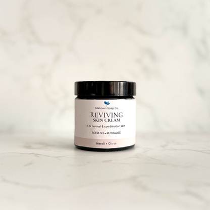 A small black cosmetic jar of Silktown Soap Co. Reviving Skin Cream with a white label, set on a light marble surface against a matching marble background.