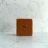 A square bar of terracotta-colored soap with a small leaf imprint in the center, standing upright on a white marble surface against a matching marble background.