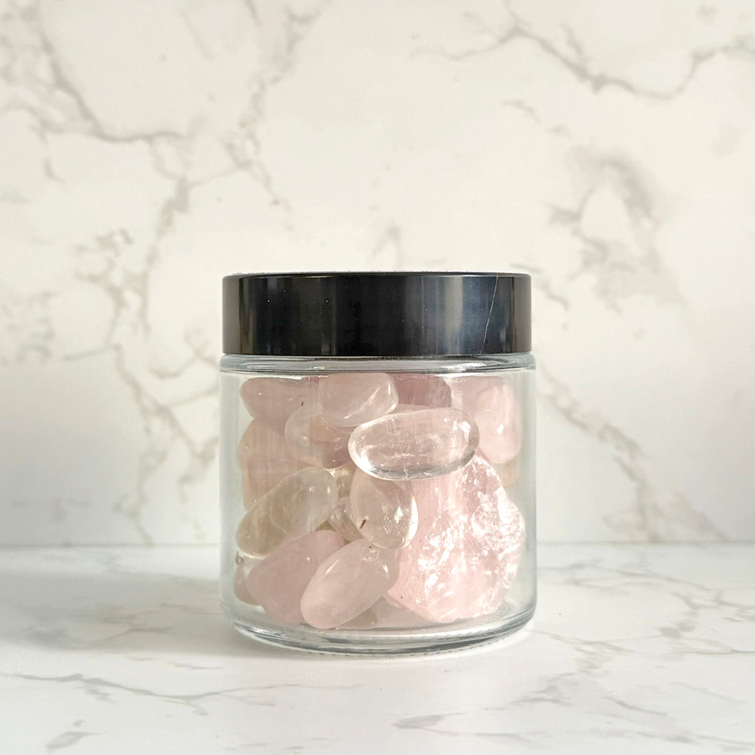 A clear glass jar with a black lid filled with smooth, polished pink tumbled stones, set against a white marble-patterned background.
