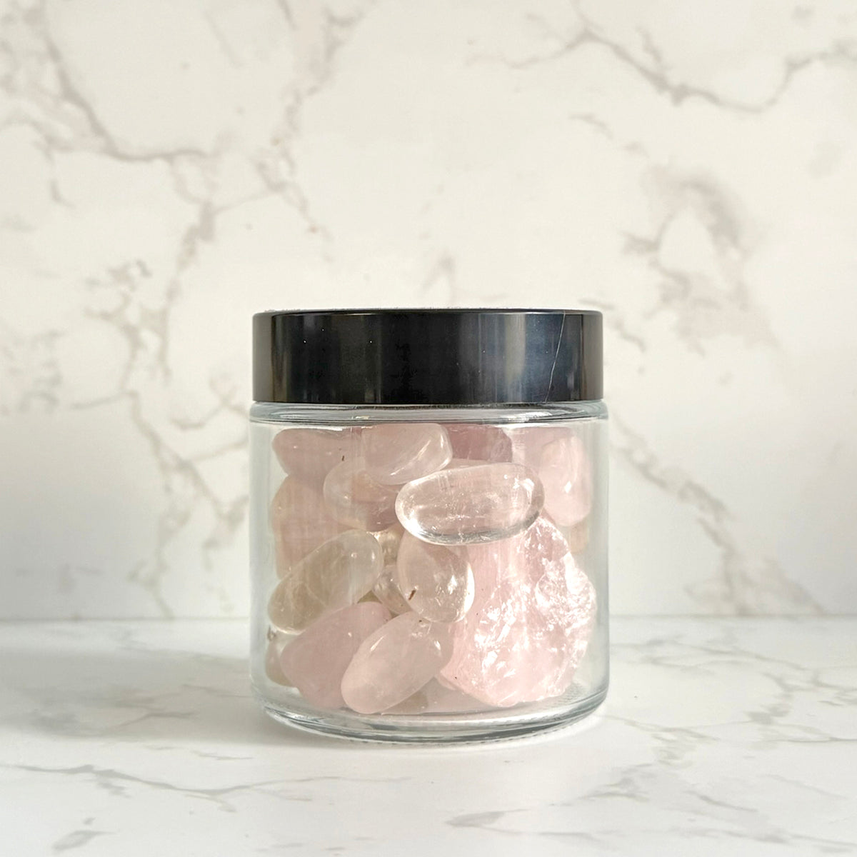 A clear glass jar with a black lid filled with smooth, polished pink tumbled stones, set against a white marble-patterned background.