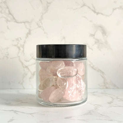 A clear glass jar with a black lid filled with smooth, polished pink tumbled stones, set against a white marble-patterned background.