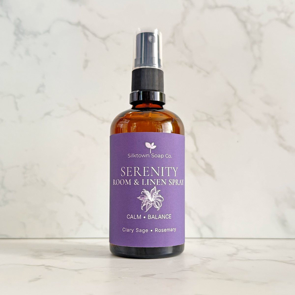 An amber glass room and linen spray bottle with a purple label from Sliktown Soap Co., standing on a light marble surface against a matching marble background.