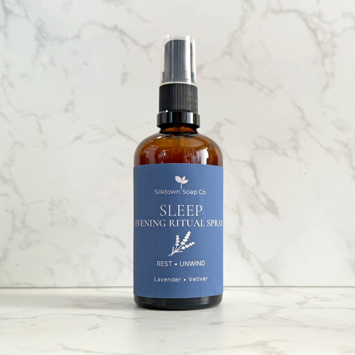 An amber glass spray bottle of Silktown Soap Co. Sleep Evening Ritual Spray with a blue label, set on a white marble surface against a matching marble background.