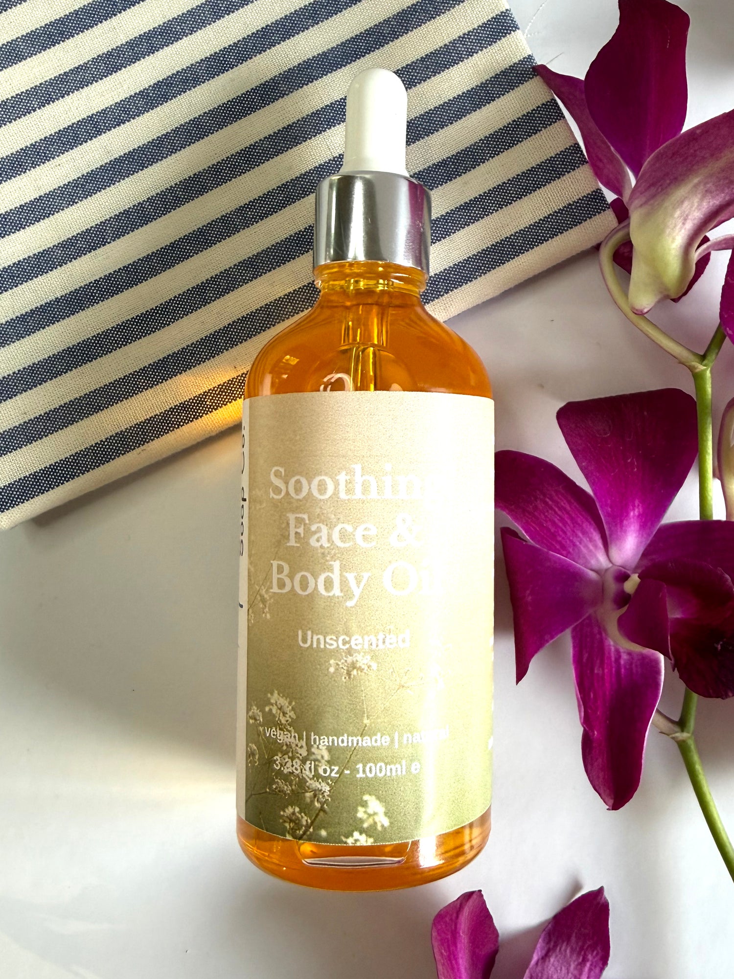 Bottle of Soothing Face &amp; Body Oil with a dropper on a white surface with purple flowers.