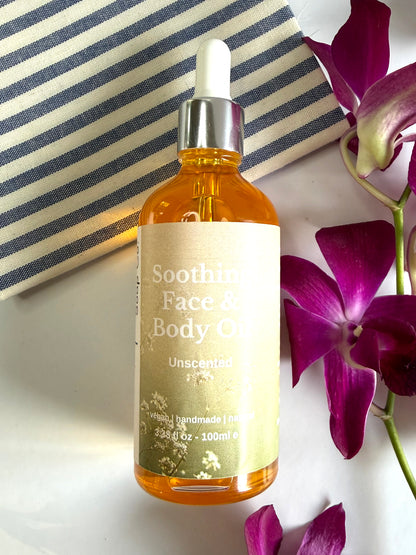Bottle of Soothing Face &amp; Body Oil with a dropper on a white surface with purple flowers.