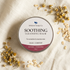 A round tin of Silktown Soap Co. Soothing Cleansing Balm, labeled Calm & Comfort, styled on a textured cream linen fabric with scattered oat flakes and chamomile flowers.