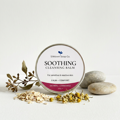 Soothing Cleansing balom tin on a white background surrounded by stones chamomile oats and olive branch