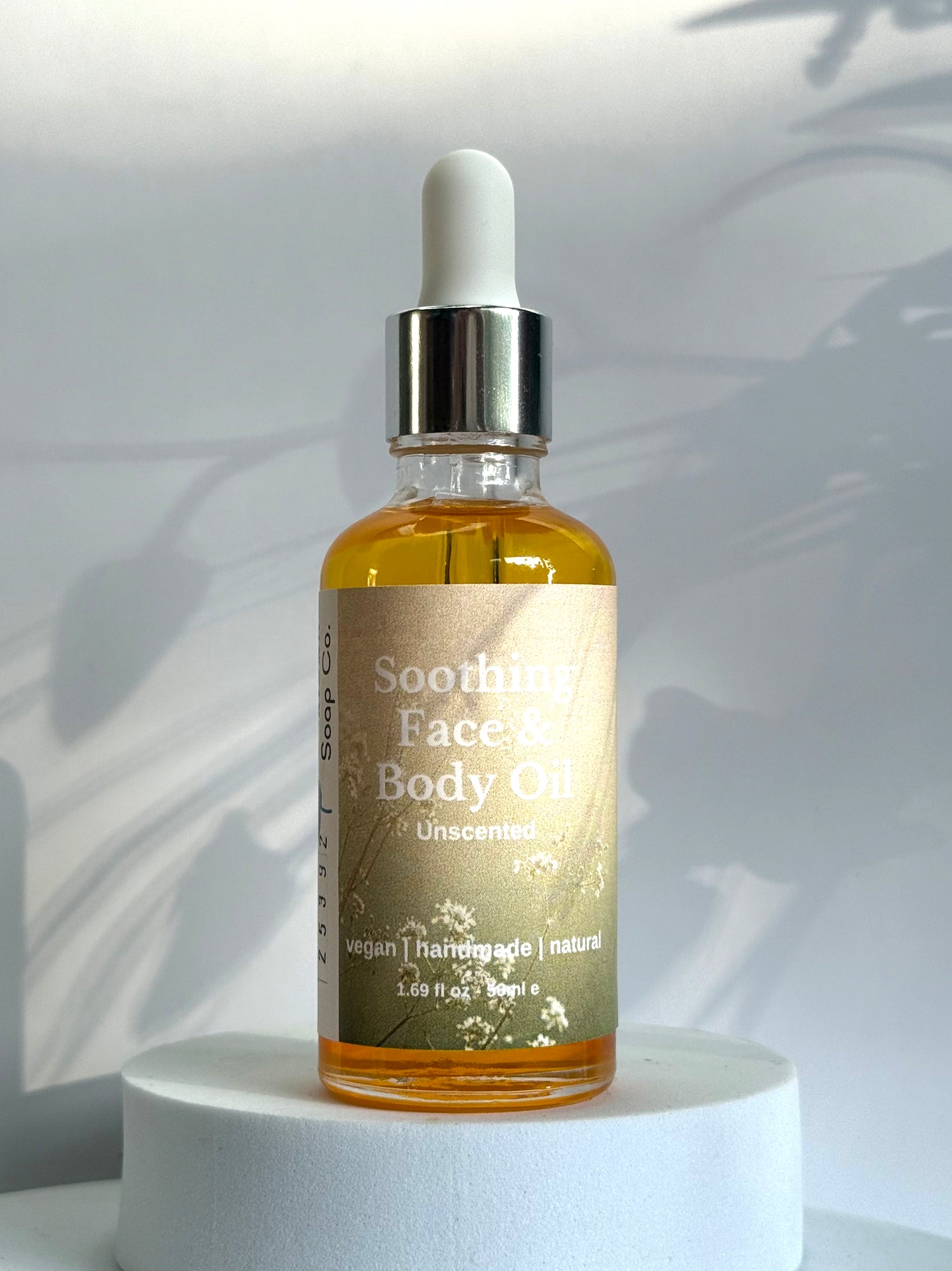 Bottle of Soothing Face &amp; Body Oil on a white pedestal with a light gray background