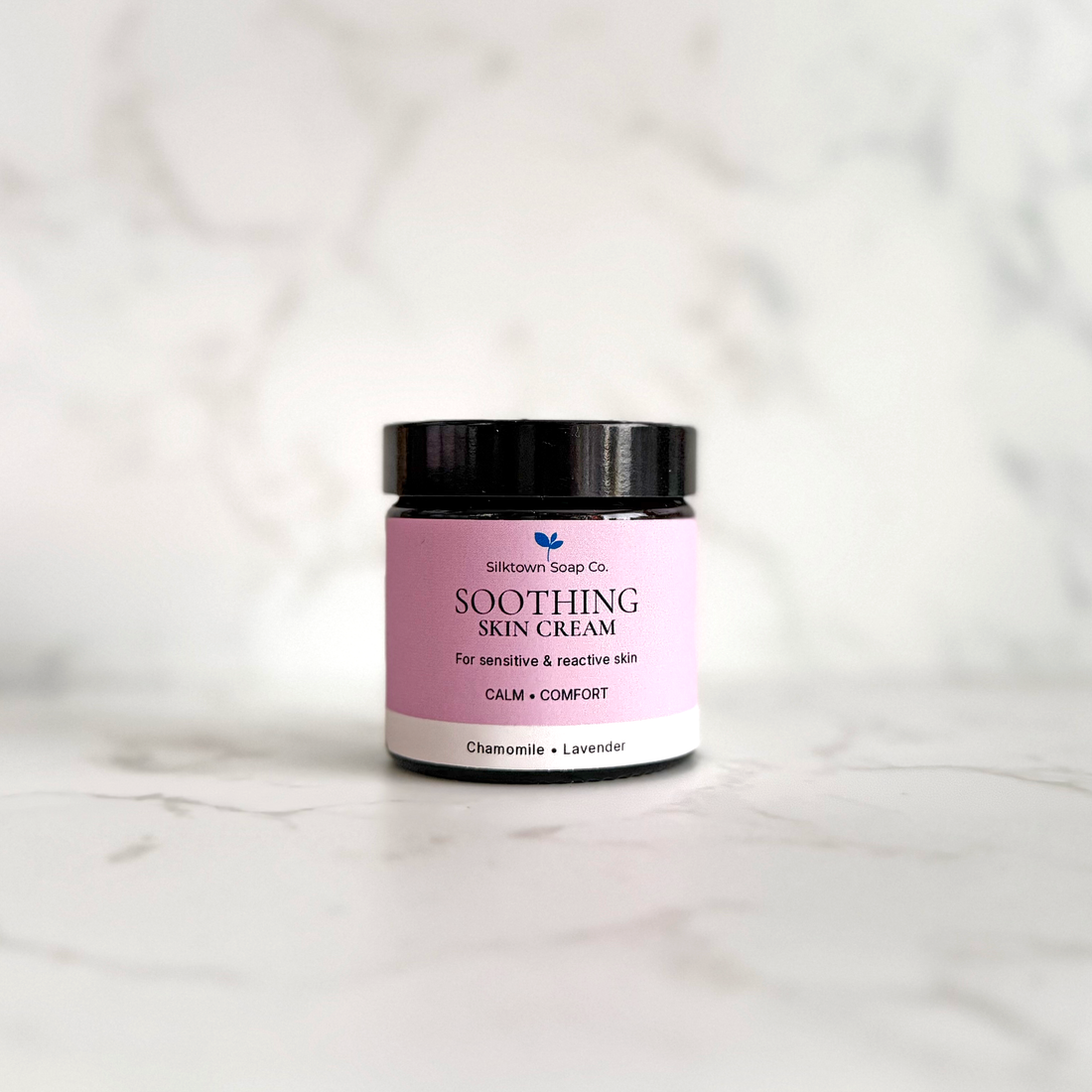 A small jar of Soothing Skin Cream by Silktown Soap Co. with a pink label and black lid, set on a light marble surface against a matching marble background.