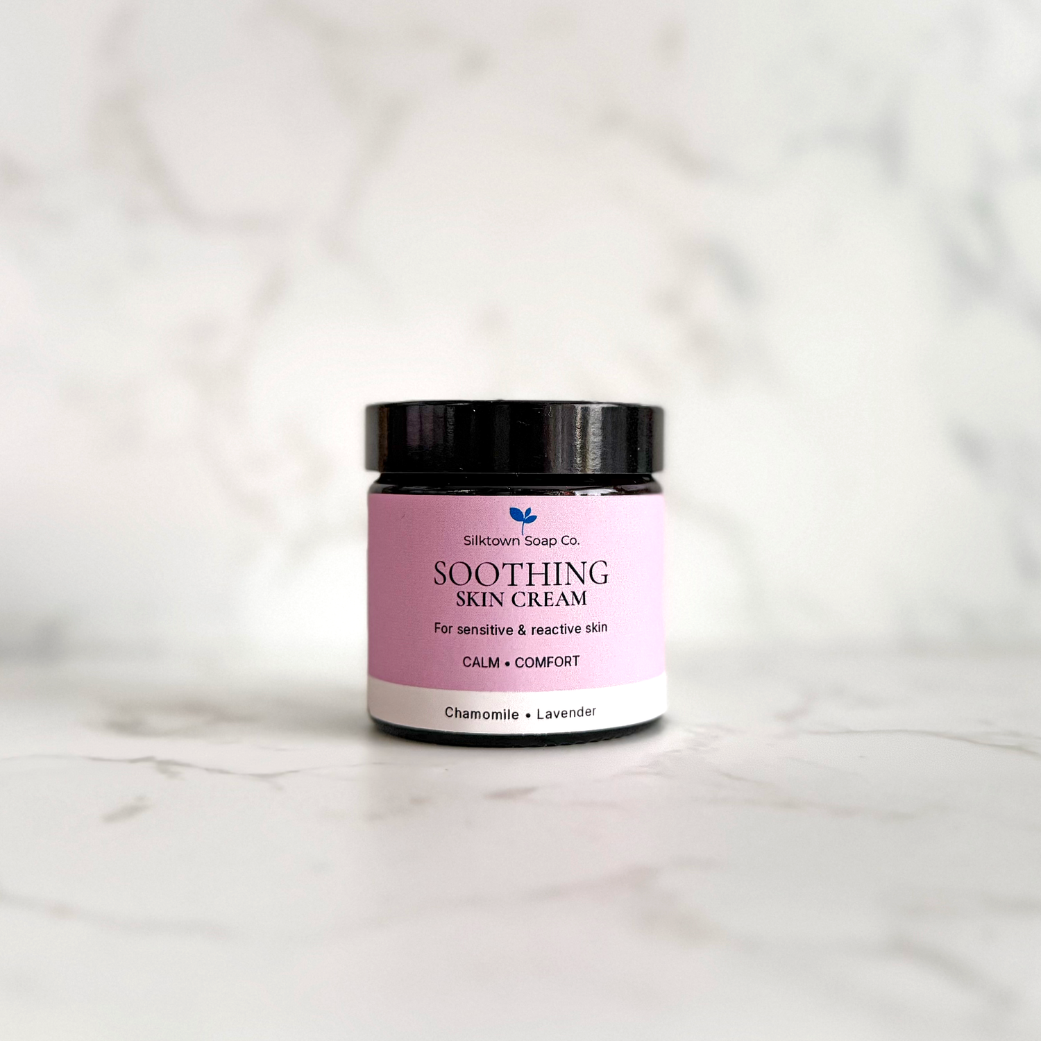 A small jar of Soothing Skin Cream by Silktown Soap Co. with a pink label and black lid, set on a light marble surface against a matching marble background.
