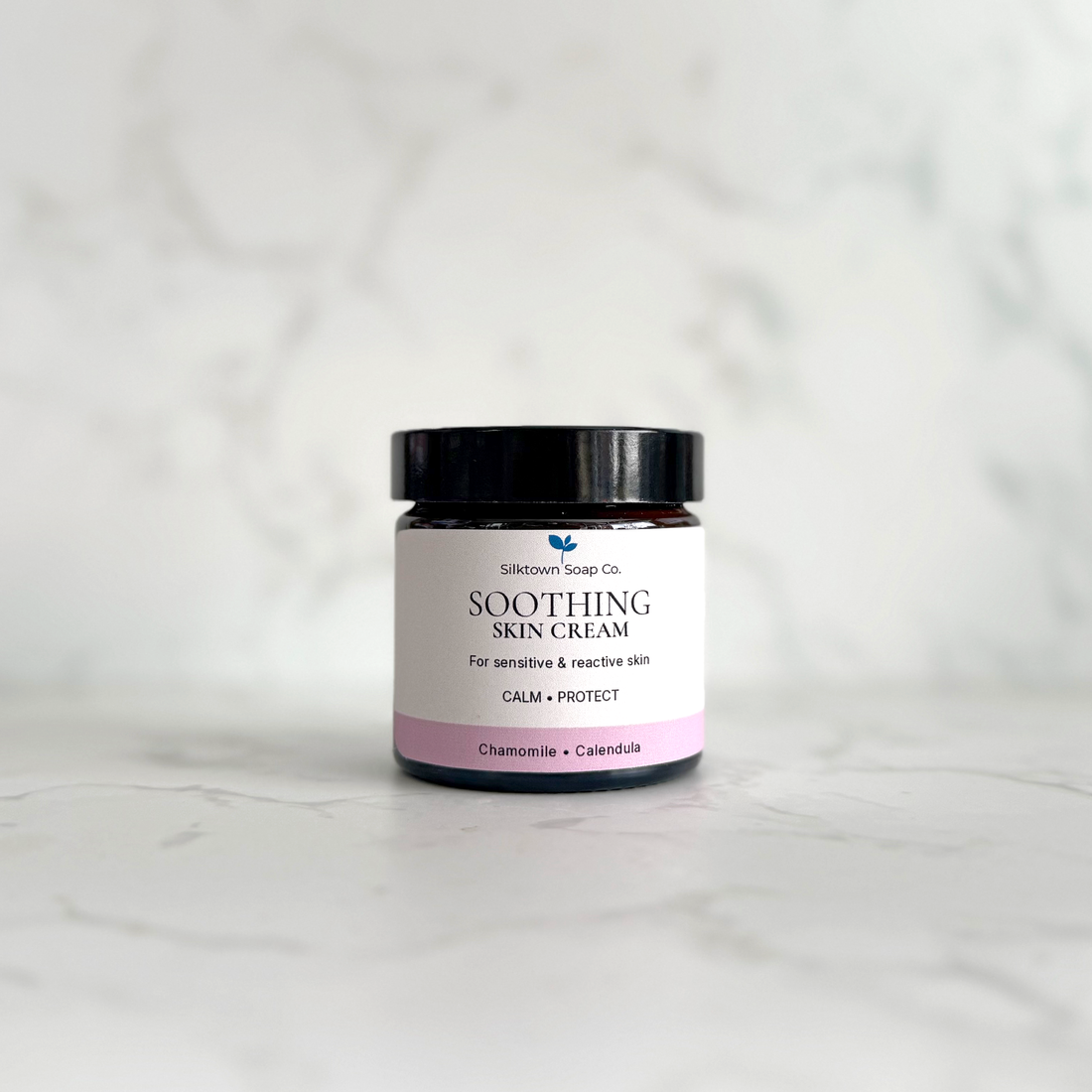 A small amber glass jar of Silktown Soap Co. Soothing Skin Cream with a black lid and a white label featuring a pink band, set on a light marble surface with a matching marble background.