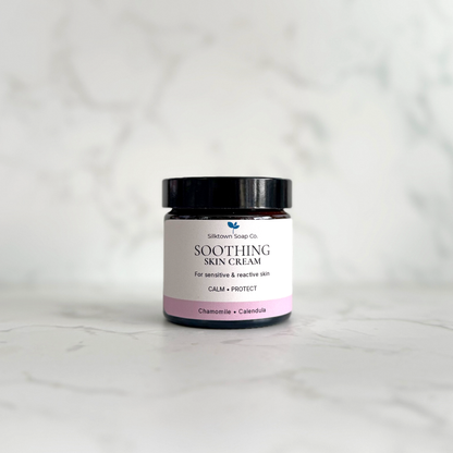 A small amber glass jar of Silktown Soap Co. Soothing Skin Cream with a black lid and a white label featuring a pink band, set on a light marble surface with a matching marble background.