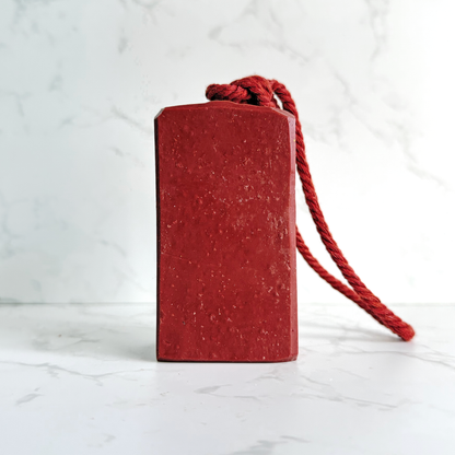 A rectangular red soap with a rounded top and a red braided cord looped through the top, standing on a light marble-like surface against a white marble background.