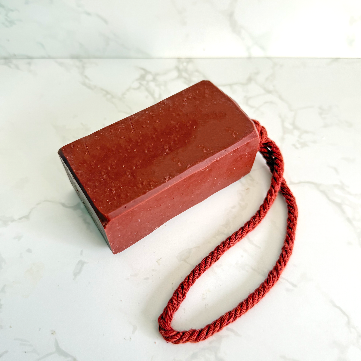 A rectangular red soap bar with a matching red twisted cord loop, photographed on a white marble surface with a white marble backsplash.