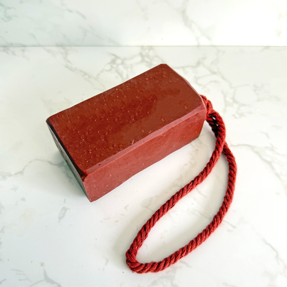 A rectangular red soap bar with a matching red twisted cord loop, photographed on a white marble surface with a white marble backsplash.