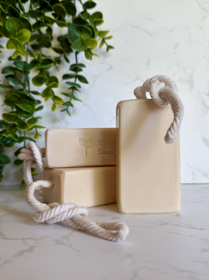 Three bars of handmade natural soap with rope handles on a light surface with greenery in the background on a marble surface