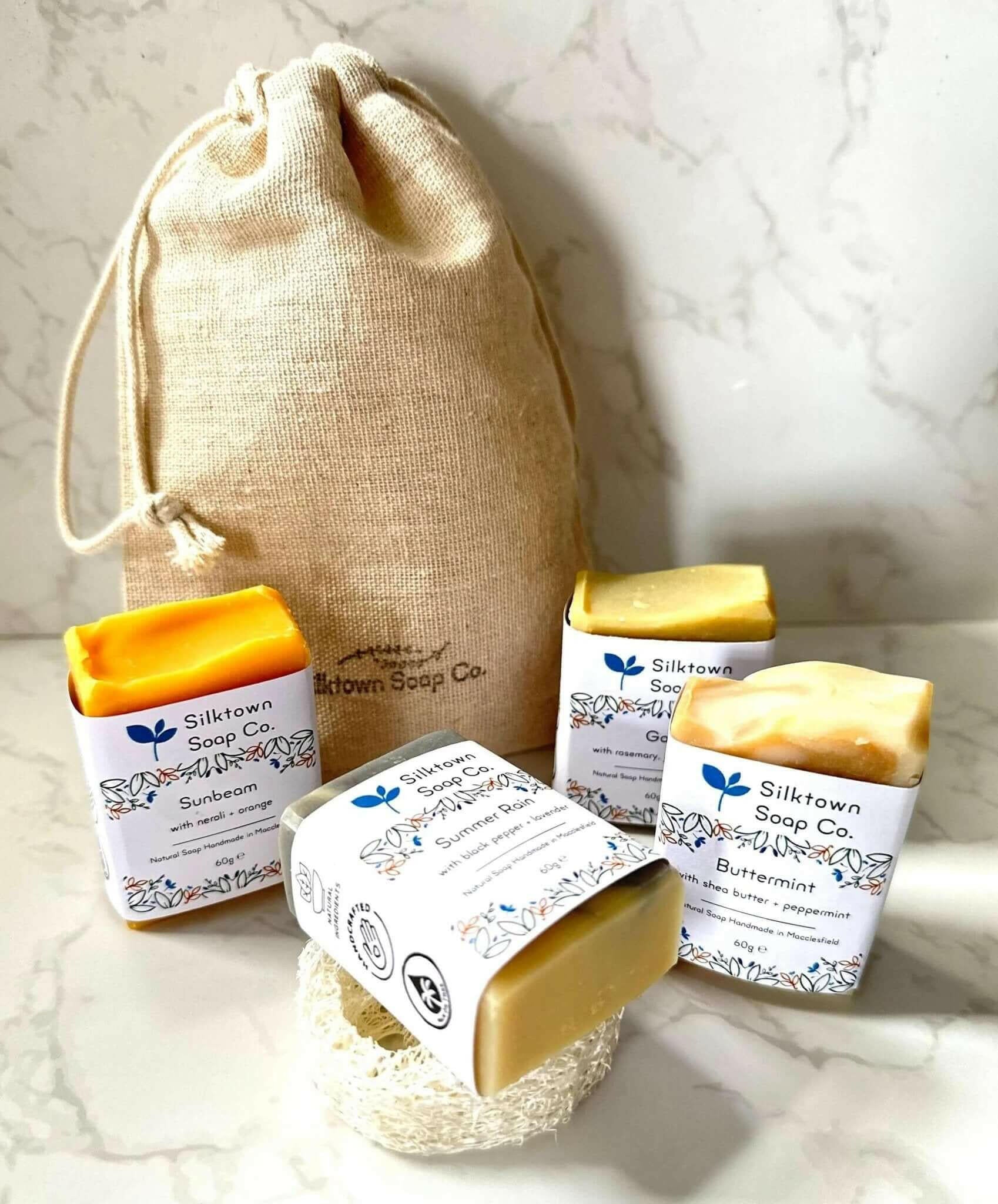 A set of four handmade natural soap, each in a different color, placed in front of a beige cotton drawstring bag, with a decorative natural loofah soap lift on the side.