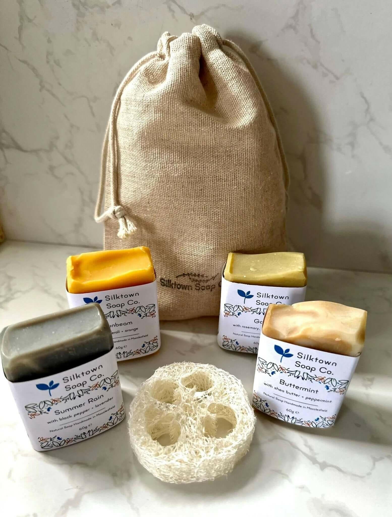 A set of four handmade natural soap, each in a different color, placed in front of a beige cotton drawstring bag, with a decorative natural loofah soap lift on the side.