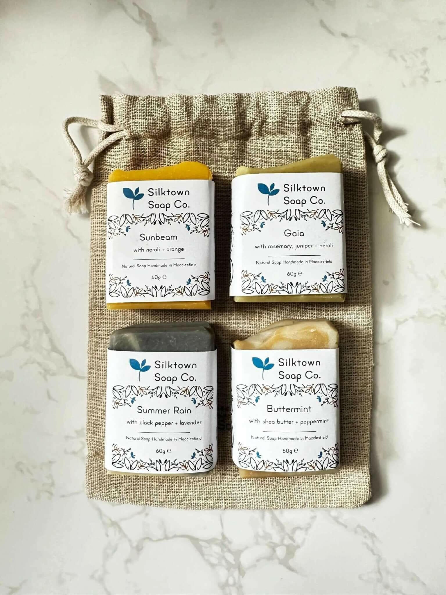 A set of four handmade natural soap, each in a different color, placed on top of a beige cotton drawstring bag.