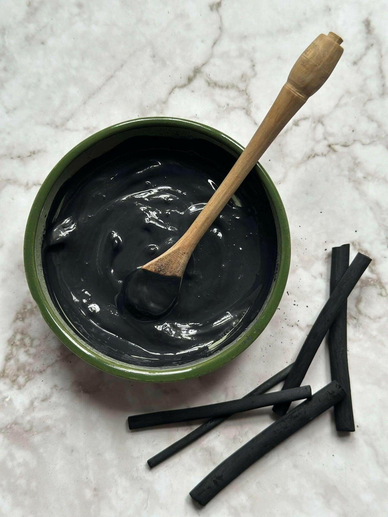 Detox Natural Clay Mask in a bowl with a wooden spoon and some natural charcoal sticks after being mixed 