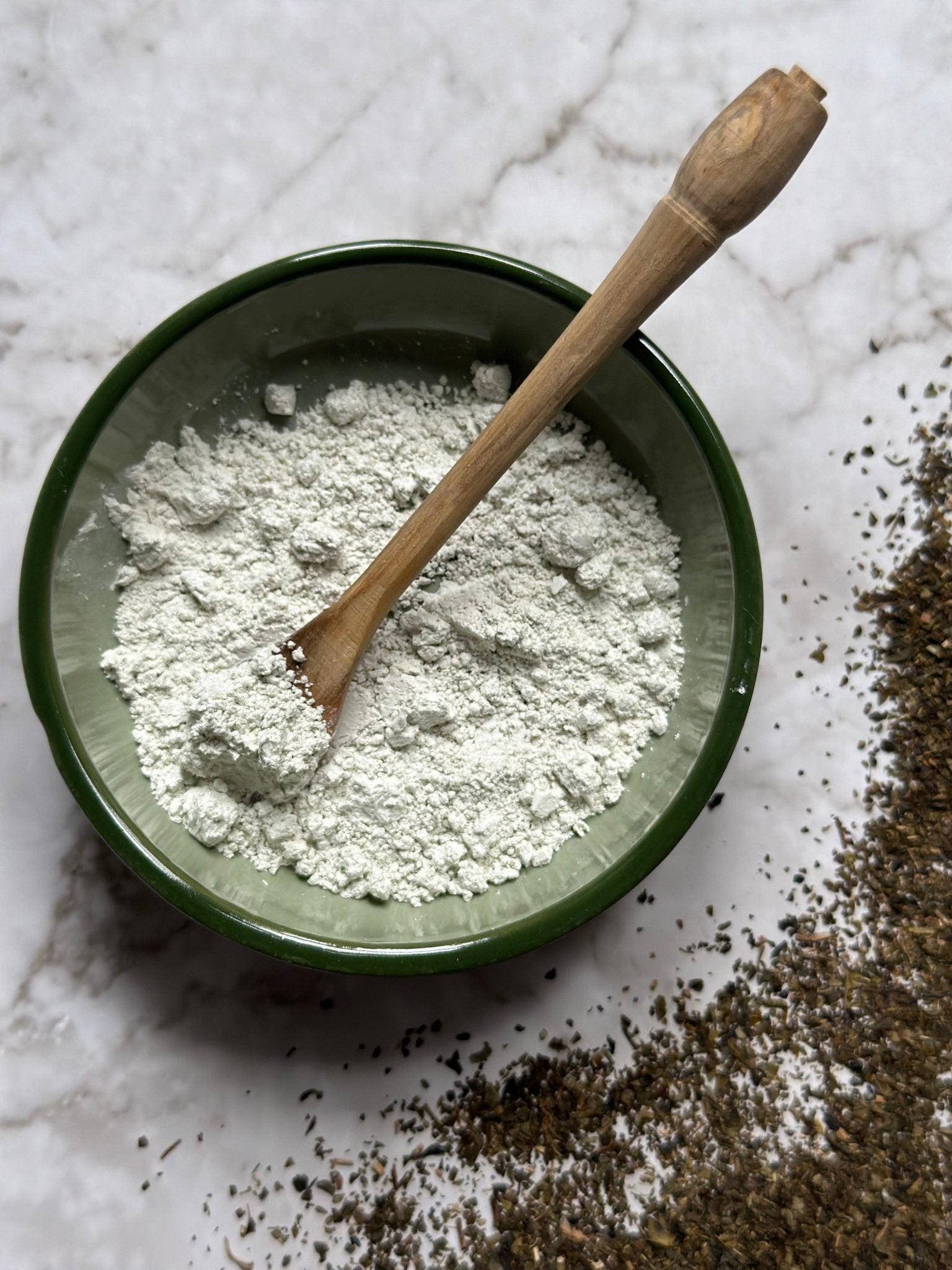 Hydrate Natural Clay Mask in a bowl with a wooden spoon before being mixed on a marble background with green tea granules